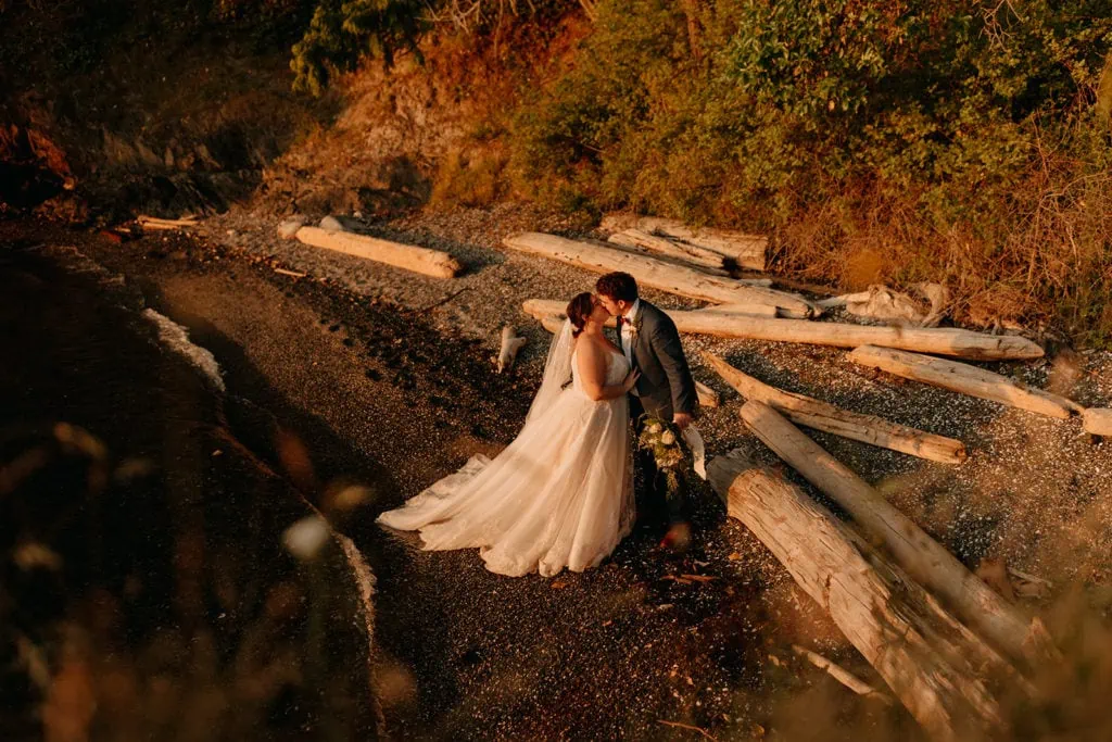 A bride and groom share a kiss in the sunset light along the shore with washed up logs near by.