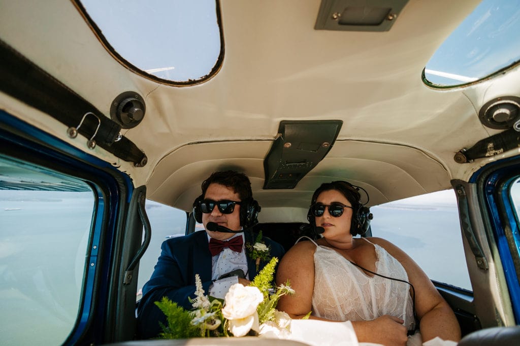 a couple sits in a seaplane with headsets and sunglasses on, fully dressed in wedding attire.