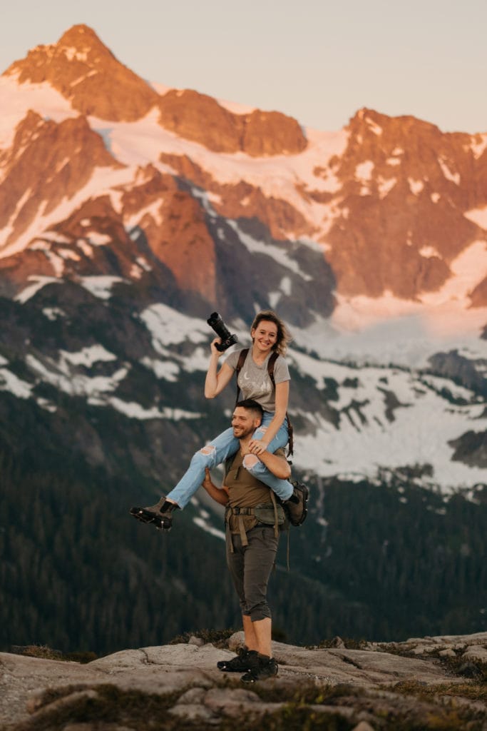 A photographer holds her camera smiling while sitting on her partners shoulders for a portrait in the mountains. 