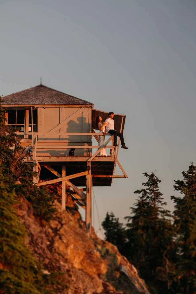 A man and woman sit on a fire tower watching the sunset.