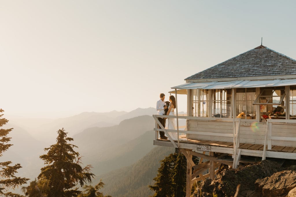 a couple holding their dog overlooking the mt baker in Washington. the fire tower is small with a shingled roof and small wooden balcony.