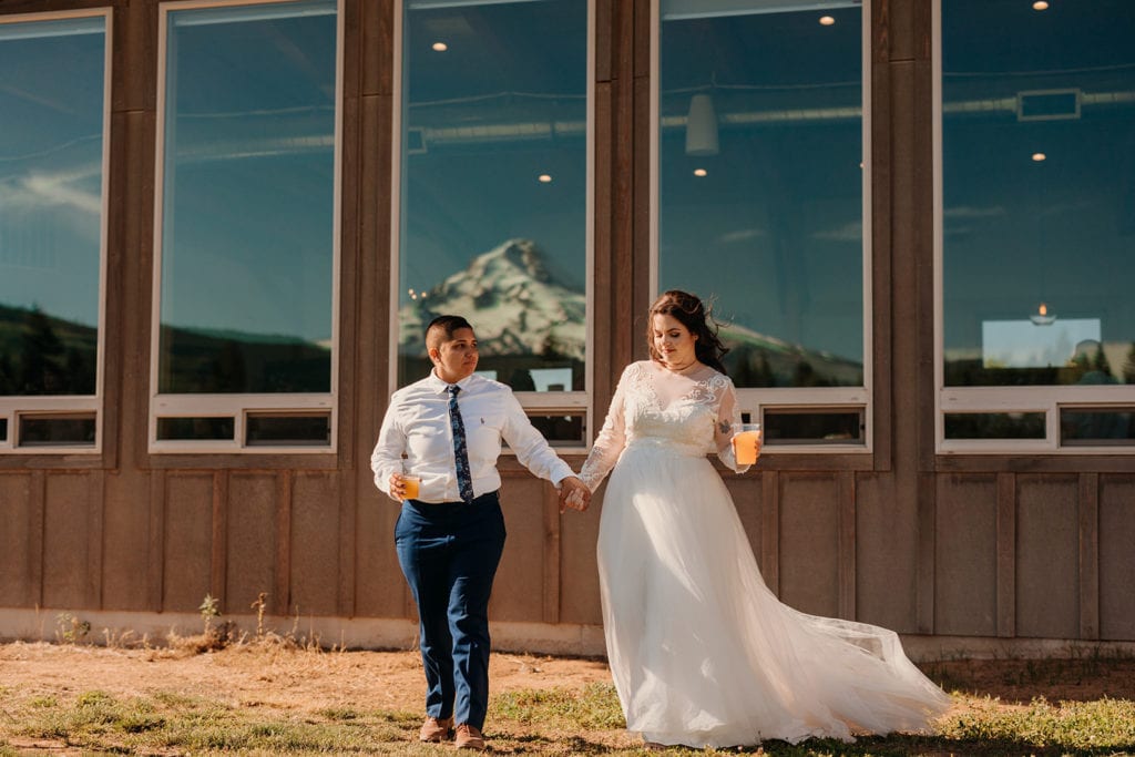 Meg and Alex hold hands and hold their beer as they look at the Mountain which is reflected in the glass behind them. 