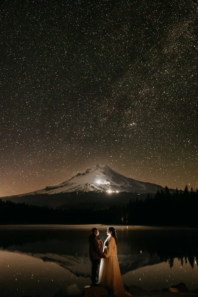 A couple stands under the milky way by a mountain.