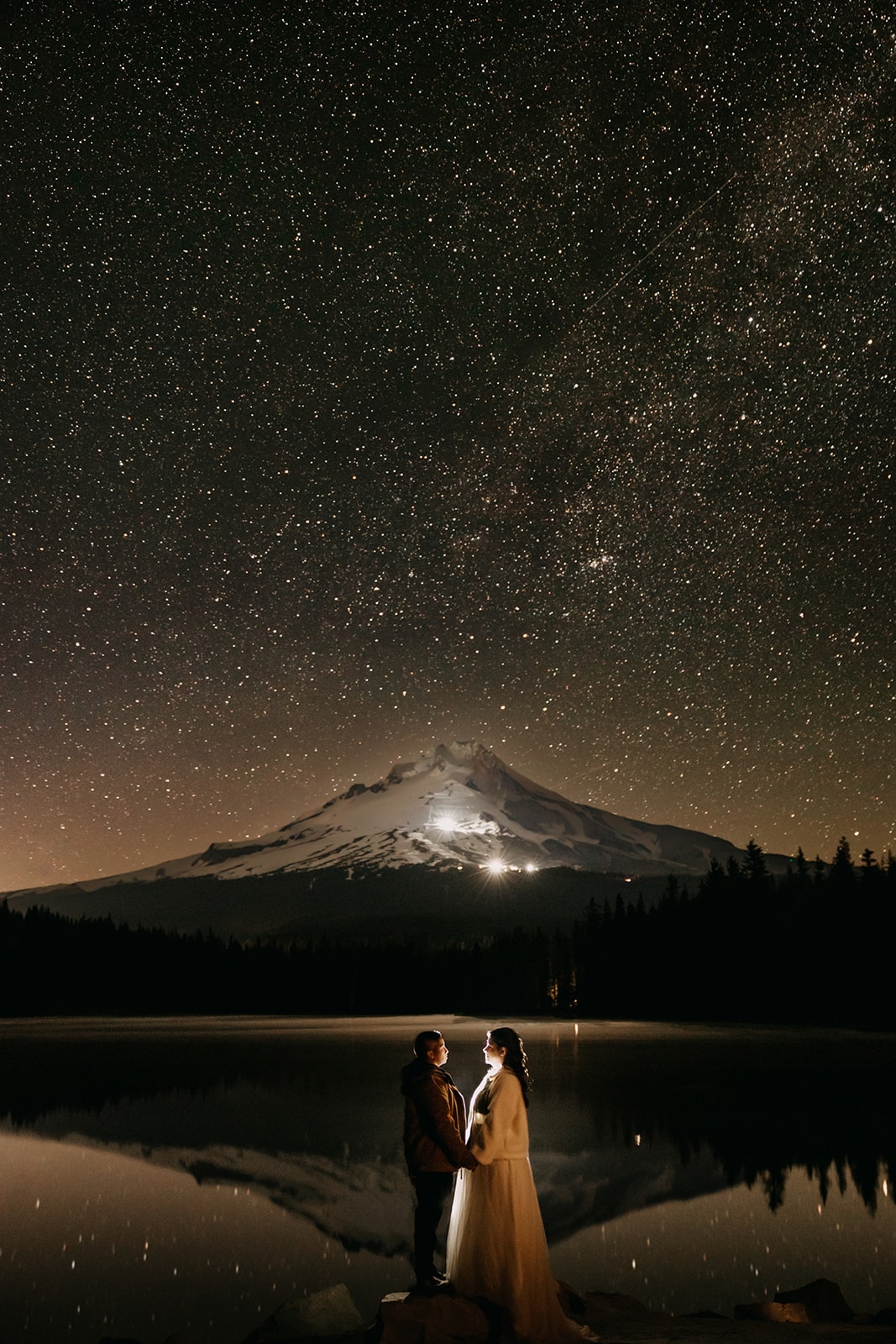 A couple stands under the milky way by a mountain.