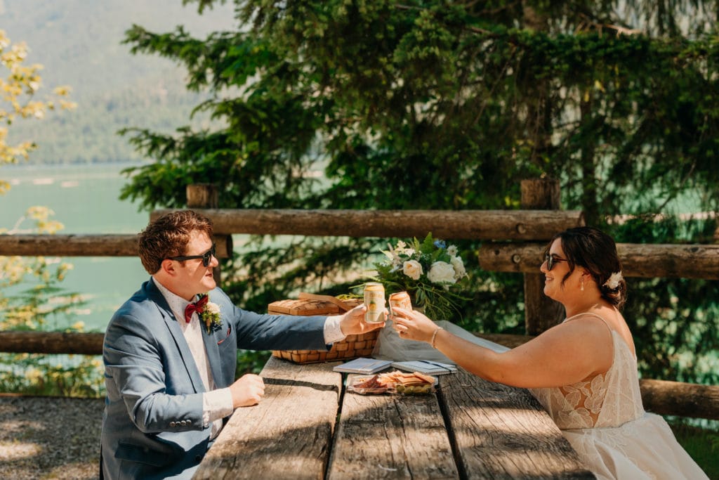 A bride and groom cheers a drink together at a picnic table.