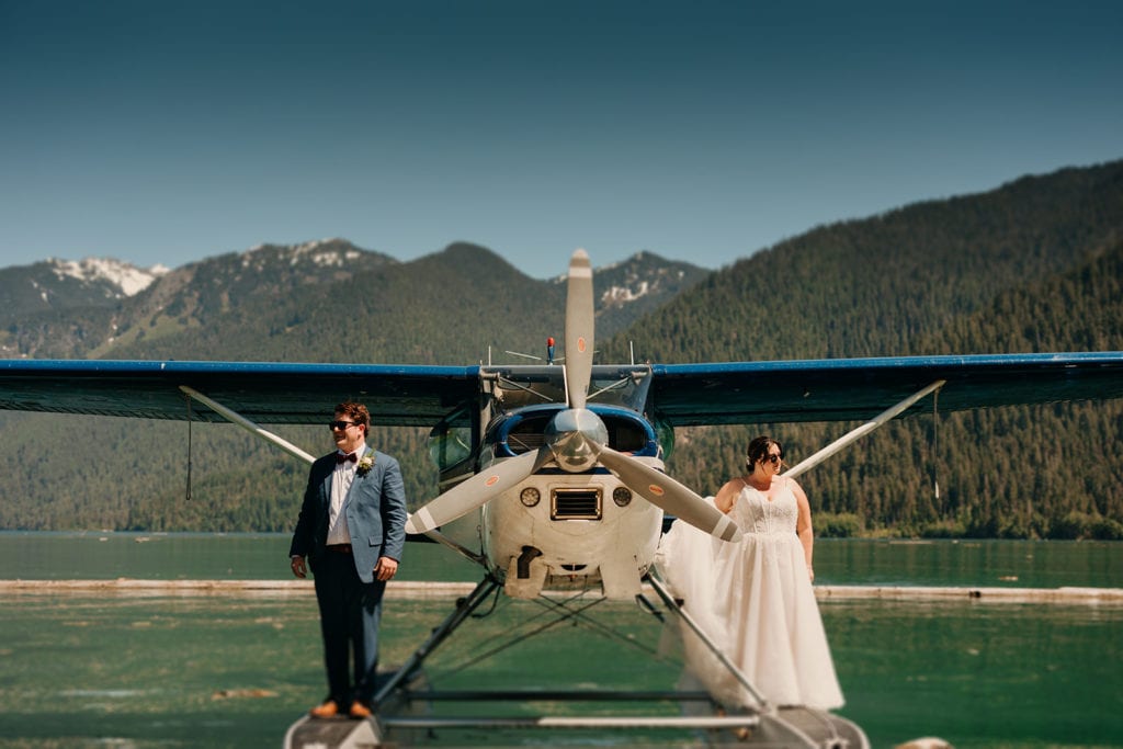 A couple stands on a seaplane on a lake in their wedding attire in the mountains. 