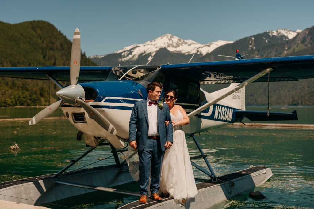 the bride holds onto the arm of the groom, smiling. they stand on the ede of the seaplane, with snowy mountains in the background.