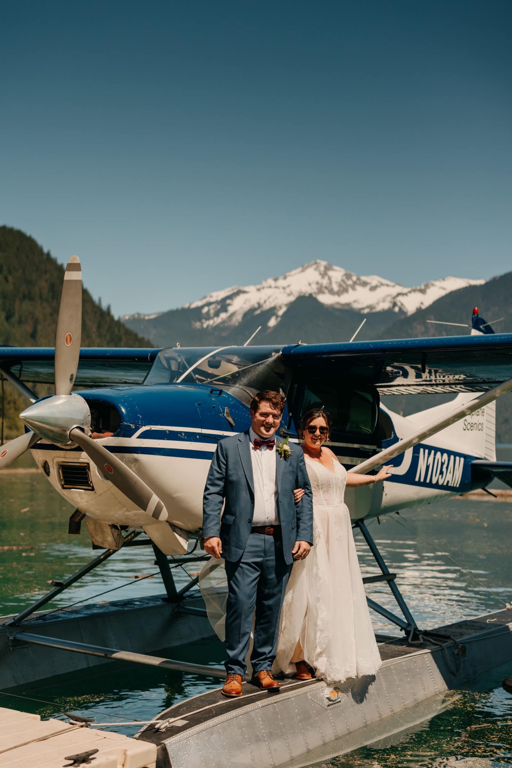 a couple stand in front of a seaplane. he wears a blue suit; she wears a white dress and sunglasses. a snowy mountain is in the background.