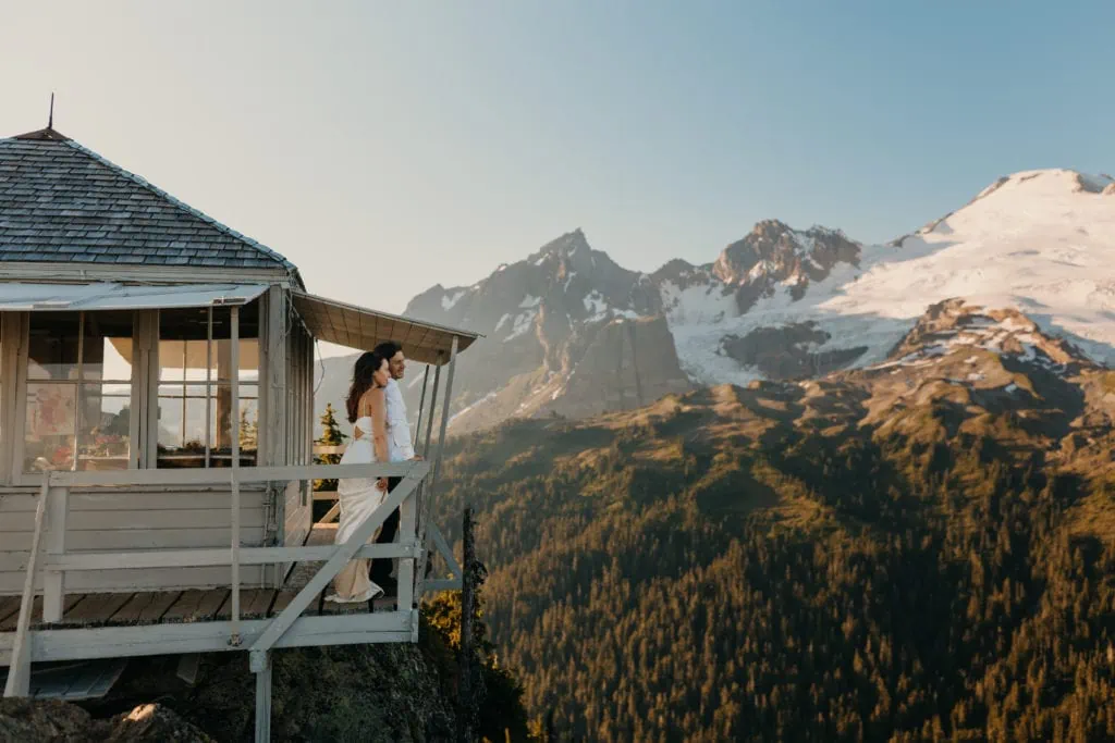 a couple overlooking the mountains on a wooden balcony of a fire tower. the man wears a white shirt; the woman wears a white wedding dress.