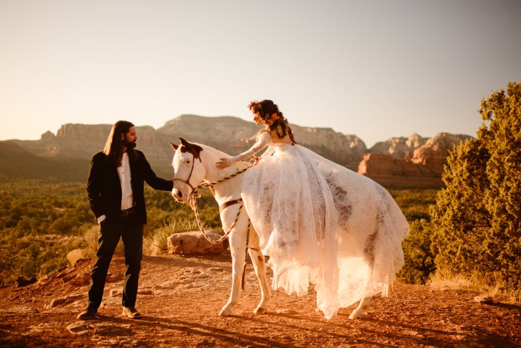 A bride pets a horse that she is sitting on in her wedding dress as the groom leads the horse. 
