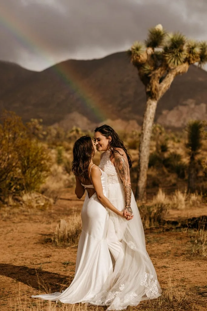 Two brides smile and hold each other close as a rainbow appears behind them at their elopement.