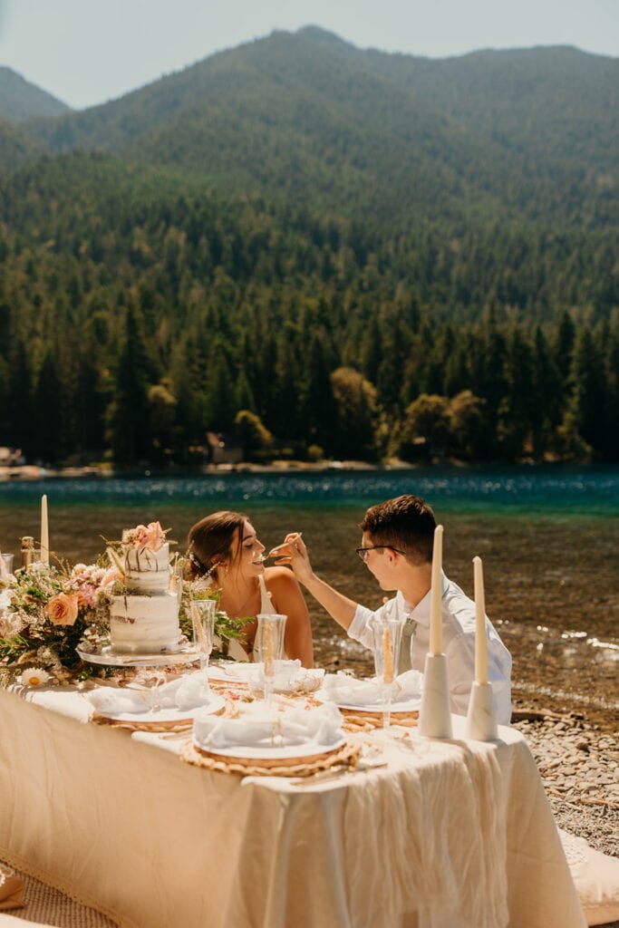 The groom feeds his bride cake at their picnic. 