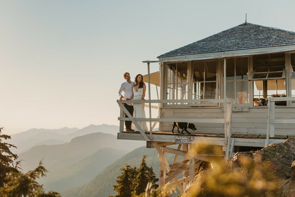 A bride and groom stand on a fire tower with their dogs nearby as they watch sunset on their wedding day.