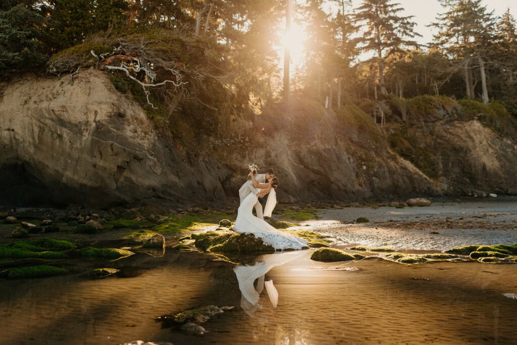 A groom dip kisses his bride as the sun rises behind them through the coastal trees. 