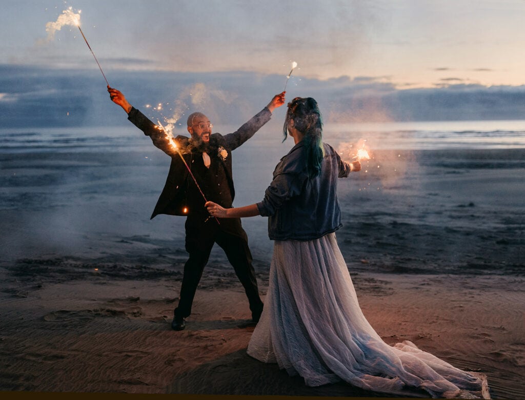 A groom showcases joy as he and his newly wed wife play with sparklers on the beach in Oregon.