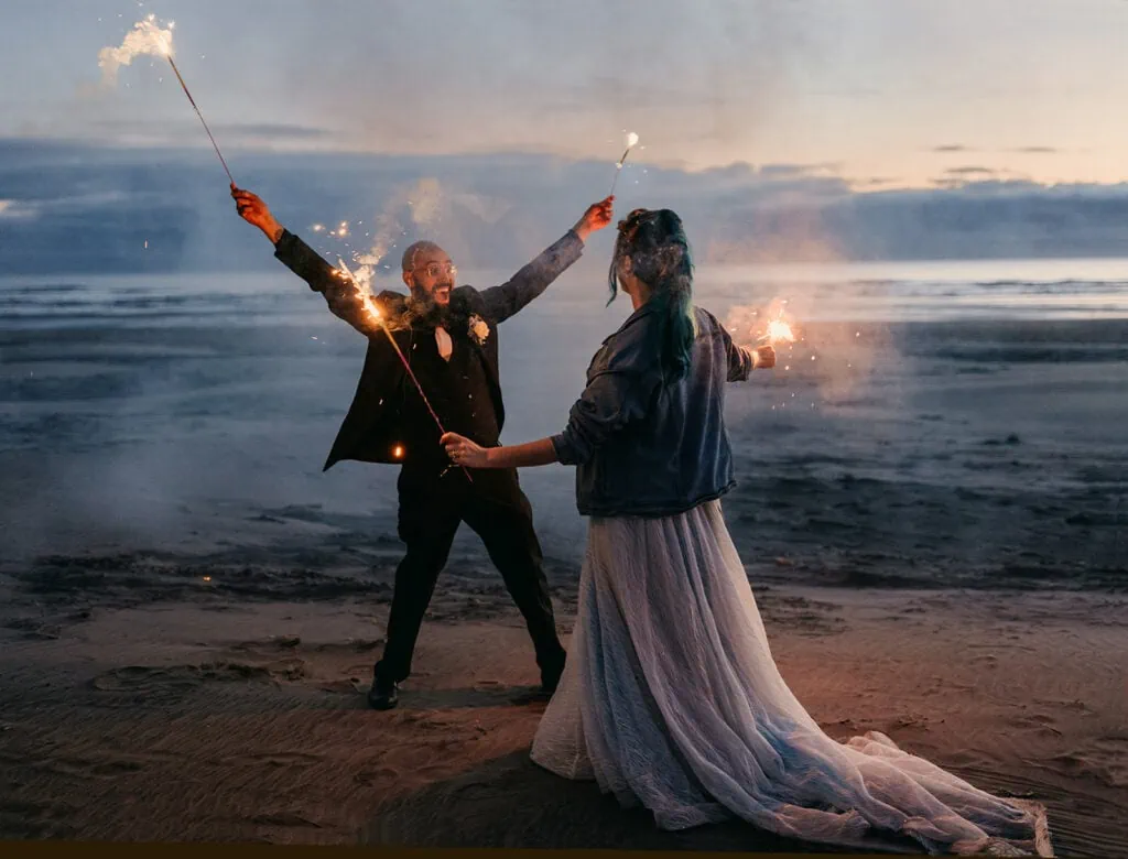 A groom showcases joy as he and his newly wed wife play with sparklers on the beach in Oregon.