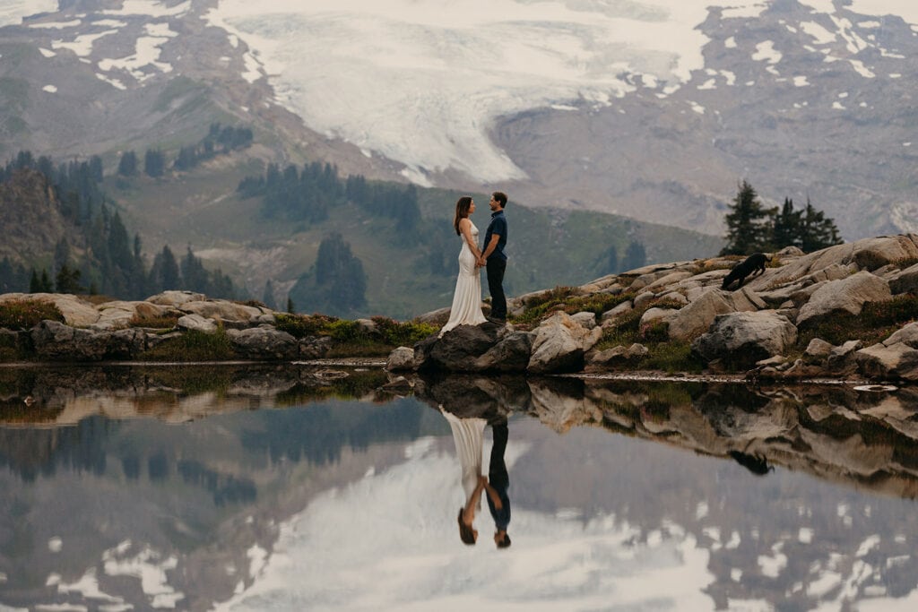 A couple holds hands and looks at each other as a glacier on a mountain stands out behind them. 