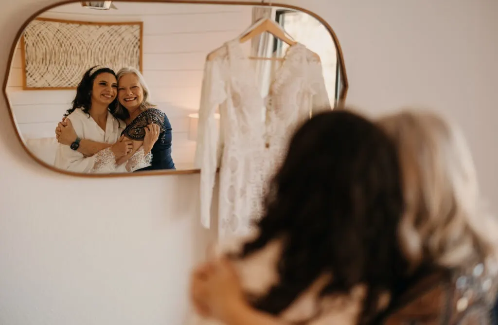 A bride and her mother look into a mirror and share a hug while the brides dress hangs on the right side of the mirror.