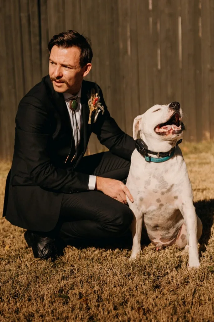 A man in a suit kneels down and pets his dog.
