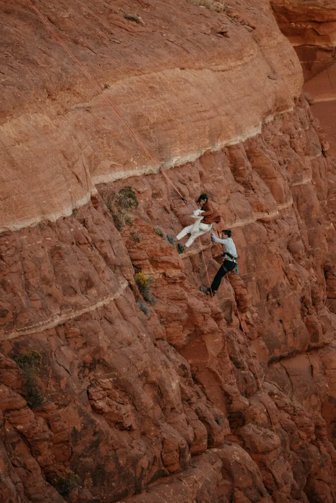 A far away shot of a bride and groom rappelling down a route they just climbed together in Sedona Arizona.