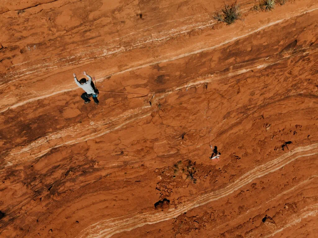 A groom climbs a red rock multi-pitch route while his bride belays him mid-day in Sedona