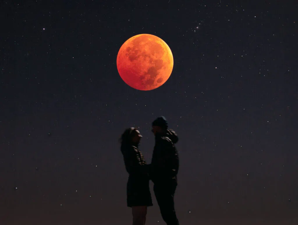 A couple stands together with the full moon above them high in the sky.