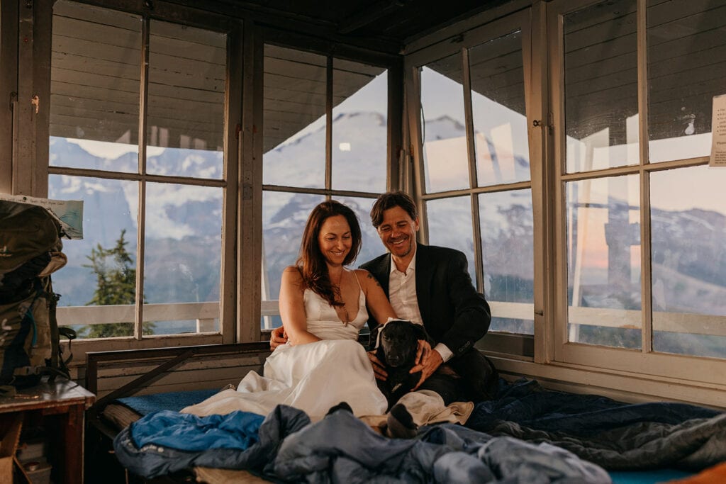 A couple smiles at their dog as they sit on a bed together in a fire tower.