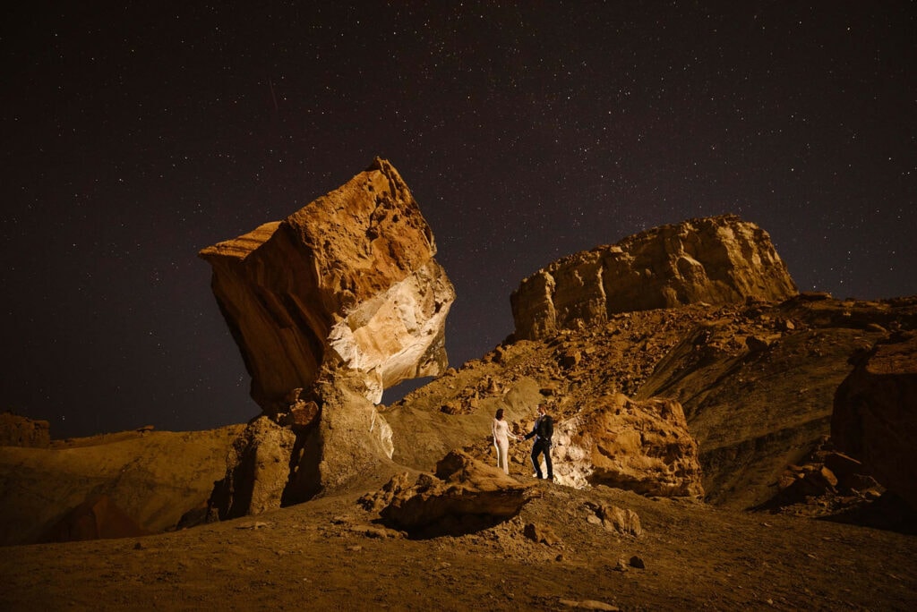 A couple stands under the stars with the lit up canyons around them.