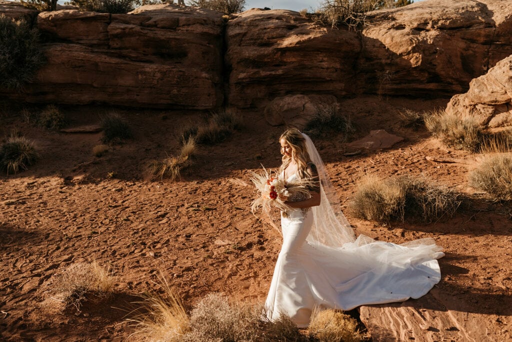 A bride walks down the trail in the sunlight towards her groom. 