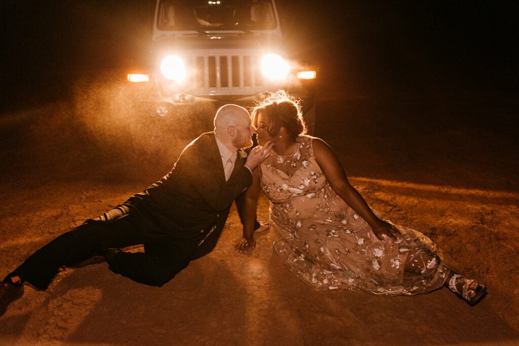 A couple kisses sitting in the dirt in front of their jeep headlights.