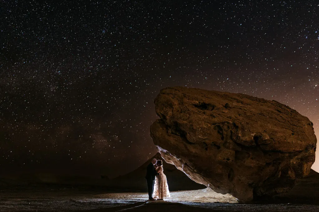 A couple stands below a boulder surrounded by the starry night sky. 