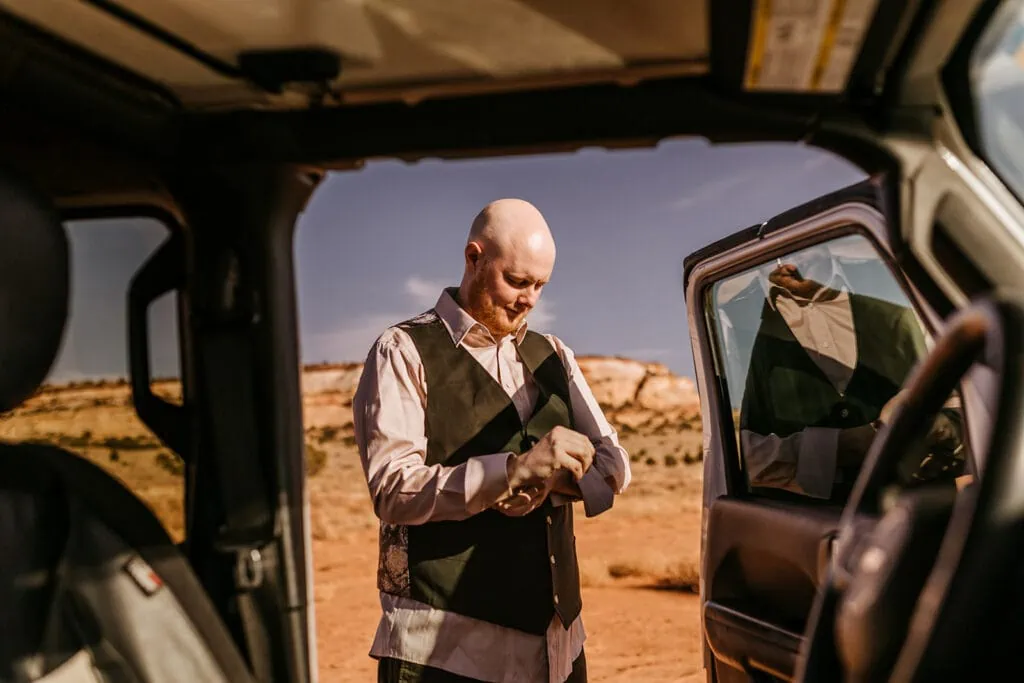 A groom puts on his suit outside of a jeep.