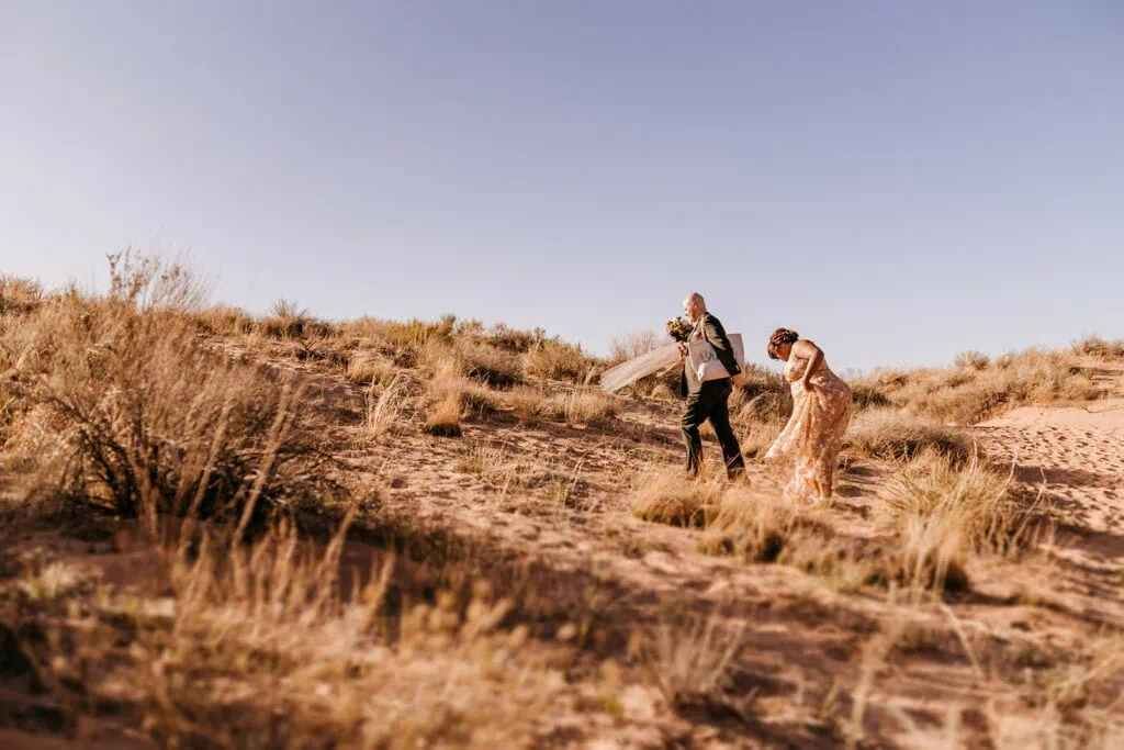 A couple walks together up the side of a sand dune.