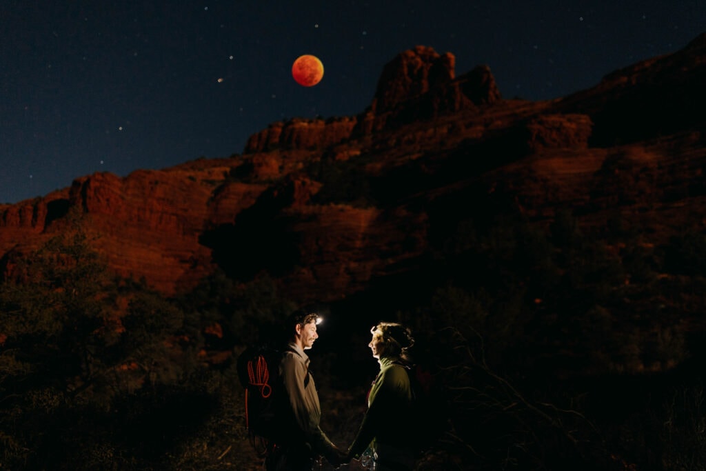 A couple holds hands in the night under the moon and red rocks.