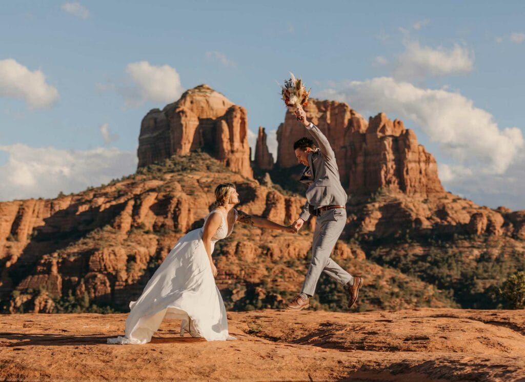 A groom jumps while holding the bouquet as the bride leans forward in the whipping wind in Sedona Arizona.