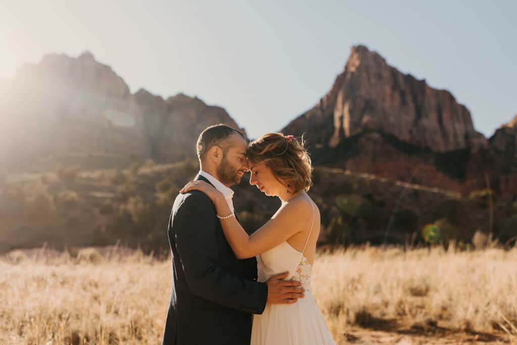 The bride and groom stand together as the sun rises in a wheat colored field among the cliffs of Zion National Park