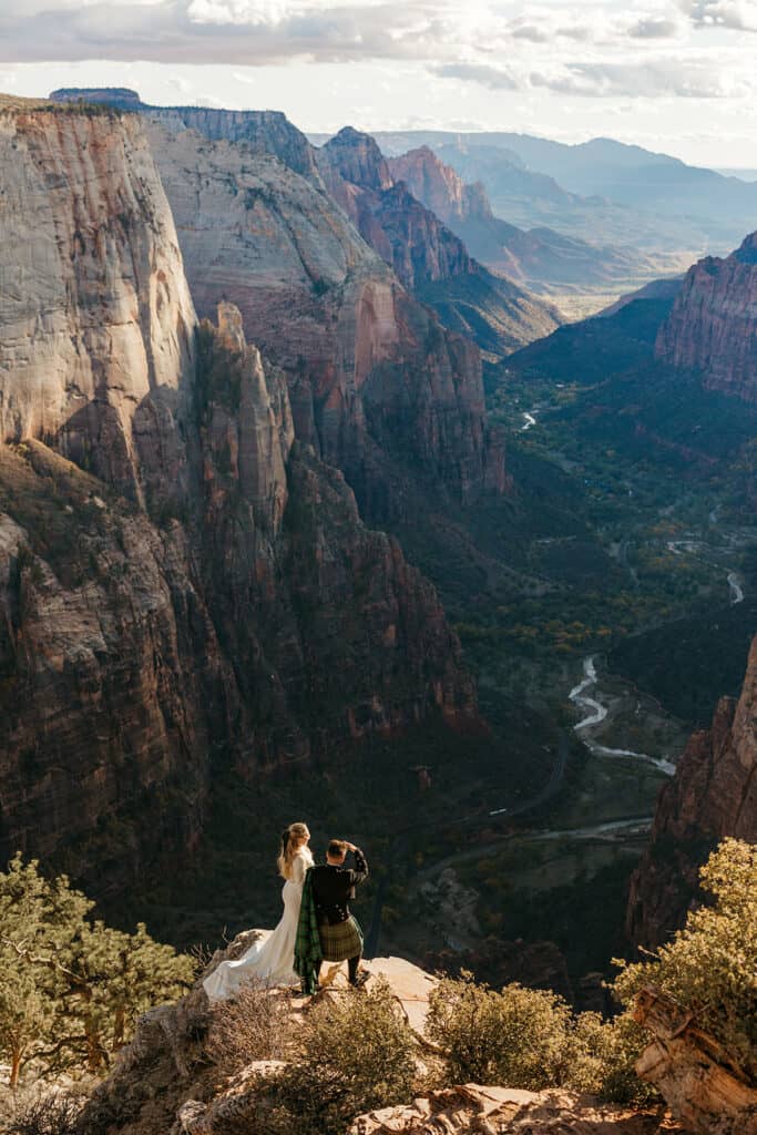 A bride and groom stand at the edge of a cliff during their elopement and take in the view down below in Zion national park.