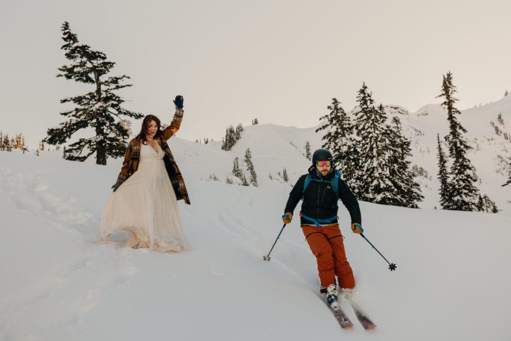 The groom skis past the bride on this snowy mountain during a winter wedding. 
