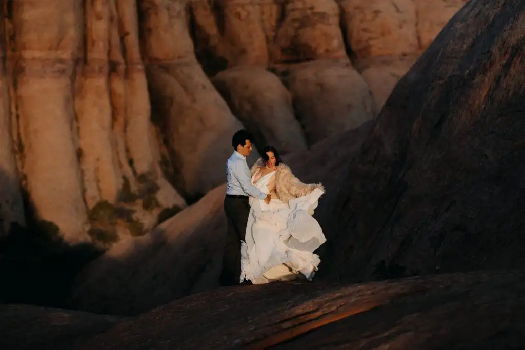 A bride plays with the fabric of her dress while in the light surrounded by a dark canyon.