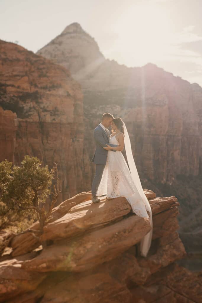 The bride and groom stand together at the edge of a cliff in Zion national park while the sun sets behind them.