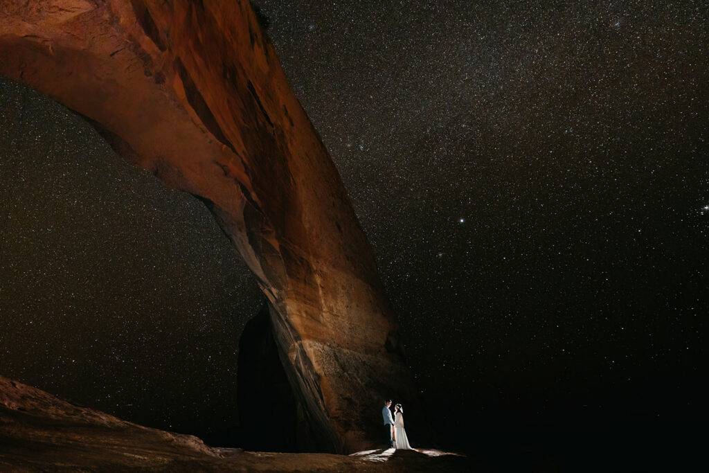 The couple stands for a portrait under the stars and a rock arch in Moab.