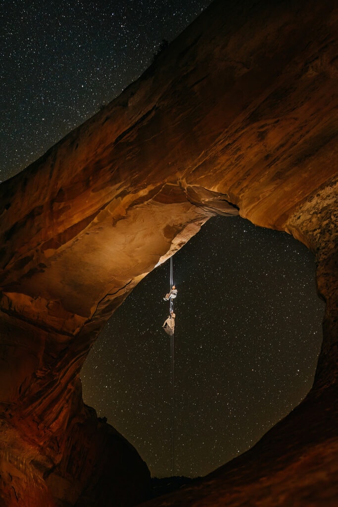 A couple hangs on rappel while star gazing from an arch.