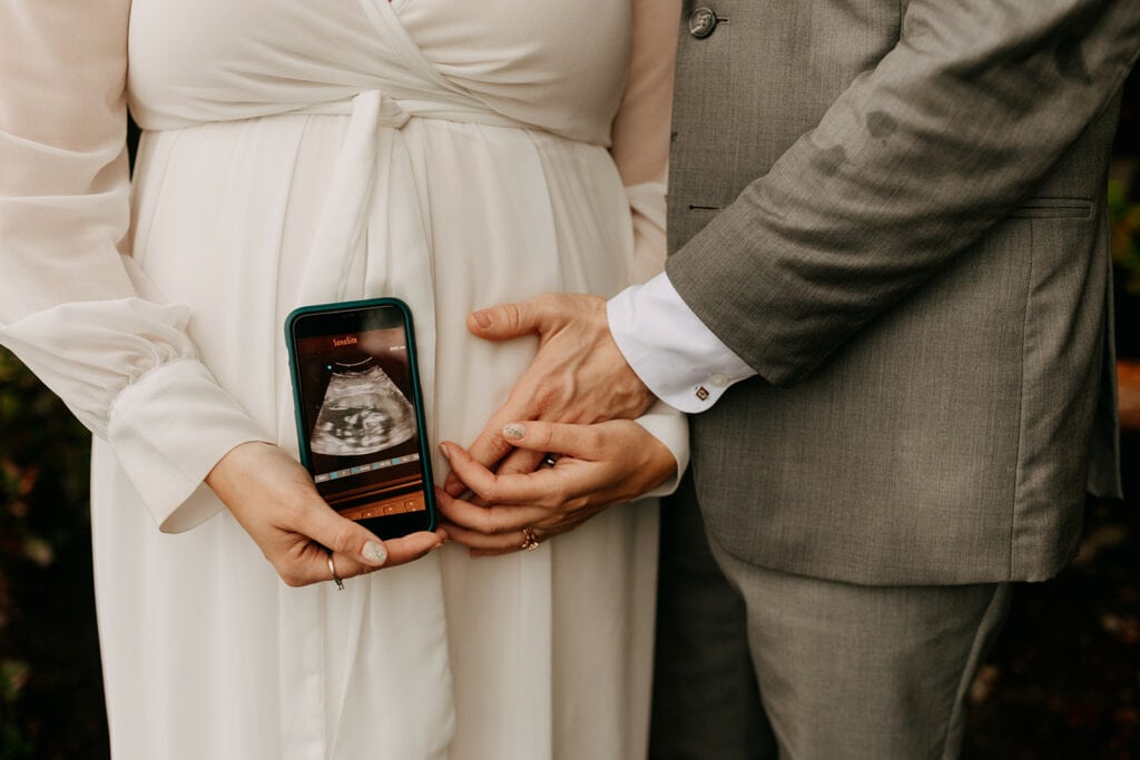 A pregnant bride and groom stand together as the groom holds the brides baby belly and the bride holds an image of her developing baby.
