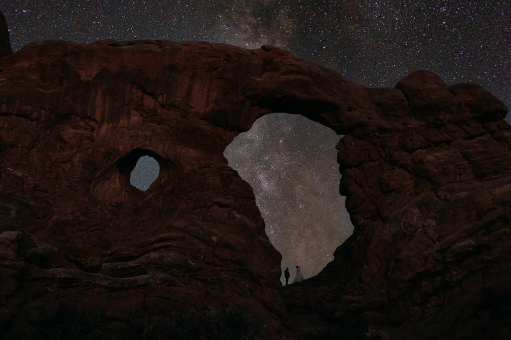 A bride and groom stand together at Arches National Park with the Milkyway passing through Turret Arch behind them