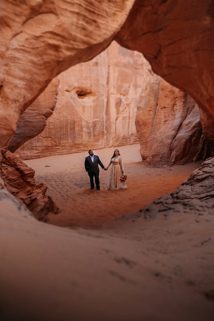 A bride and groom stand together under sand dune arch in arches national park