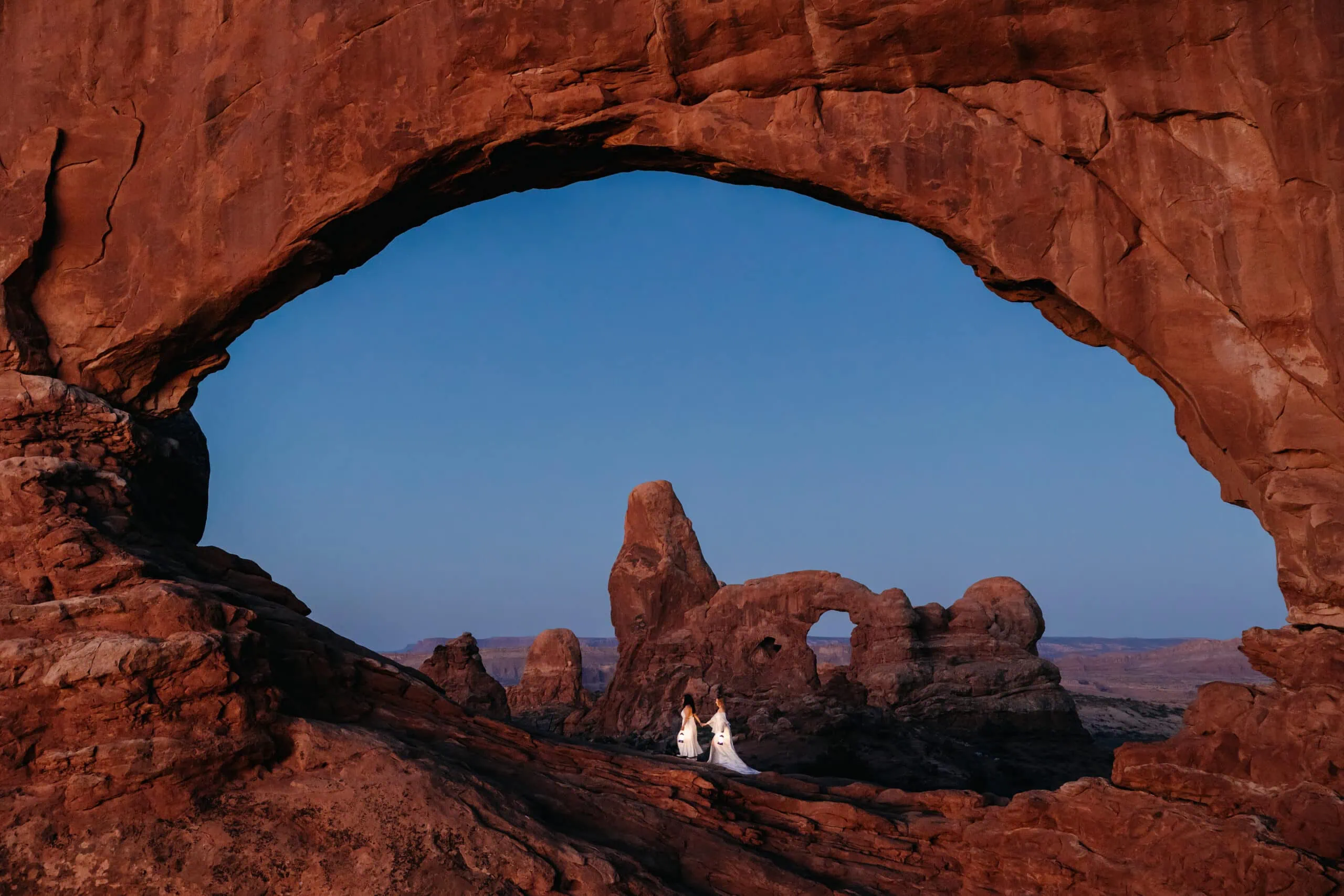 Two brides walk across North Window arch at dusk.