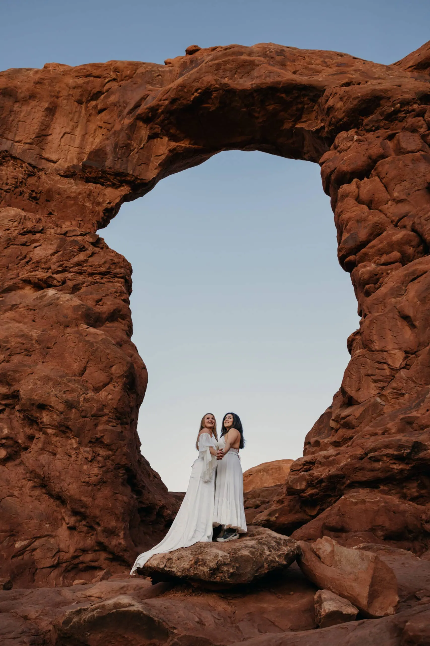 Two brides hold each other as they watch the sun peak above the horizon in Arches National Park.