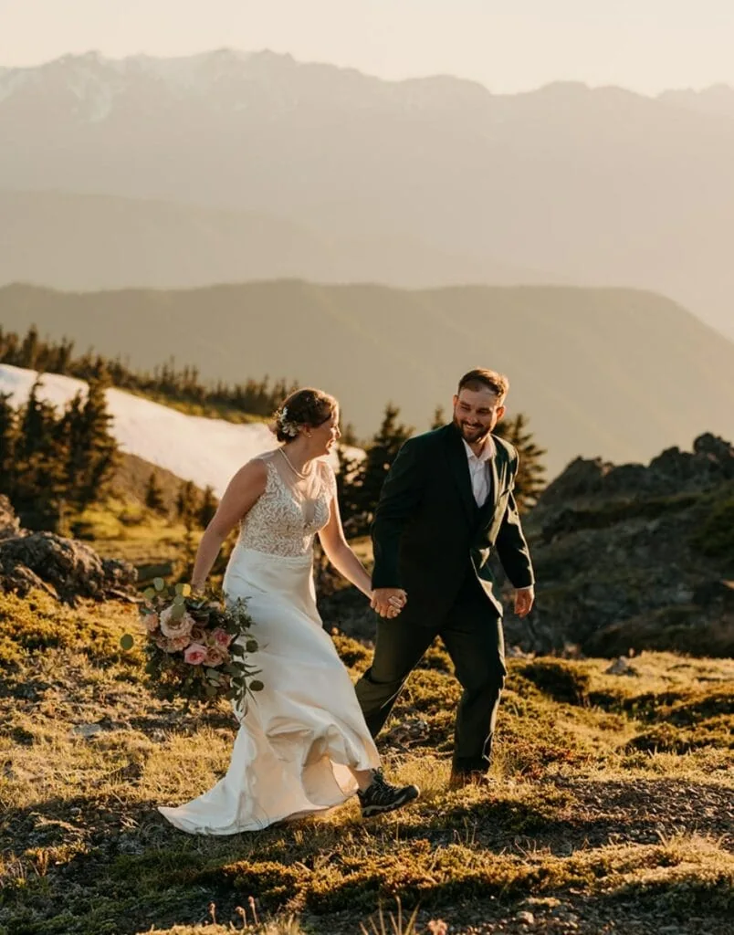A bride and groom walk together holding hands through a mountain area