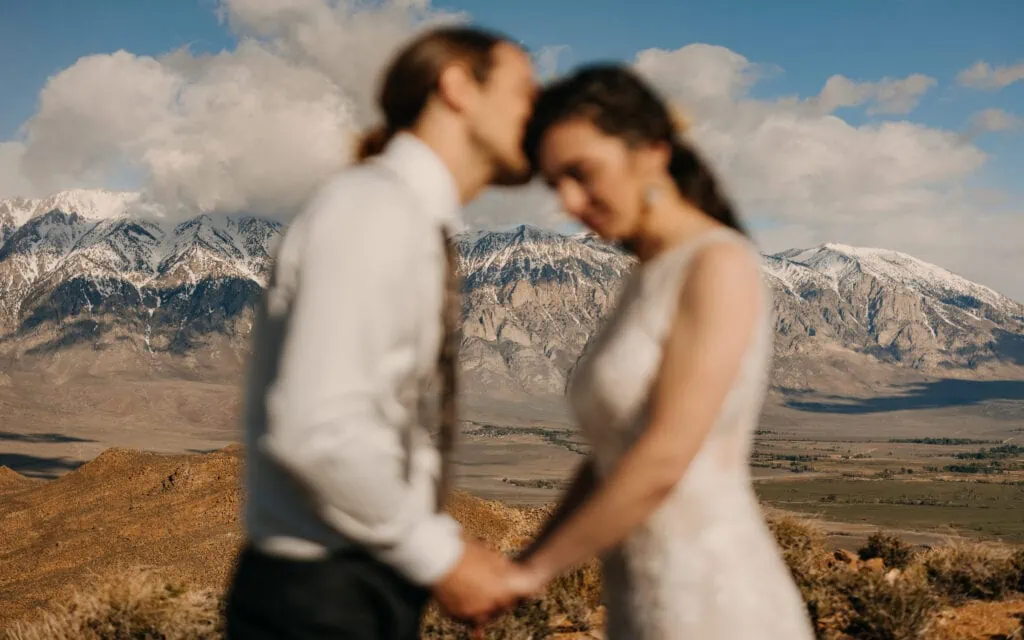 A portrait of the couple holding hands as the groom kisses the brides forehead but the focus is on the mountains behind them.
