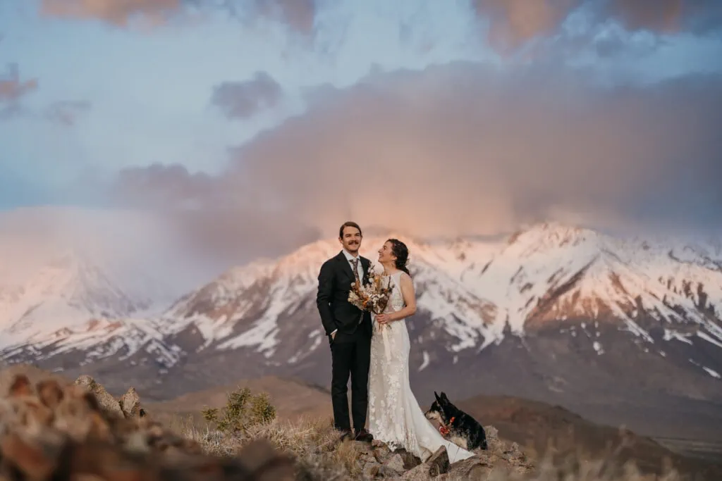 The couple smiles as they stand for a portrait at sunrise in the mountains.